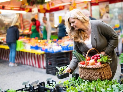 Eine Frau kauft auf einem Wochenmarkt ein und hält einen mit frischem Obst und Gemüse gefüllten Einkaufskorb in der Hand.