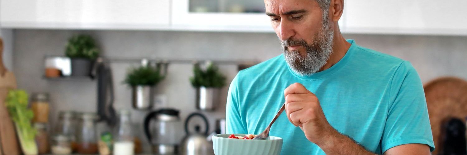 In einer Küche sitzt ein grauhaariger, bärtiger Mann im T-Shirt und isst Obst aus einer Schüssel.