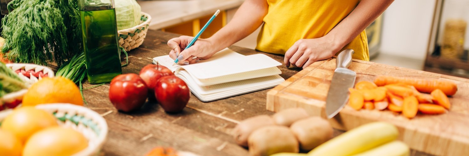 Eine Frau steht an einer Arbeitsplatte mit Gemüse und Obst und schreibt in ein Heft.