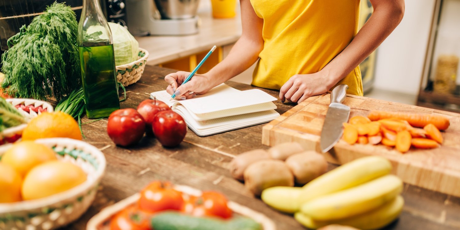 Eine Frau steht an einer Arbeitsplatte mit Gemüse und Obst und schreibt in ein Heft.. Artikel "Kochen" öffnen.