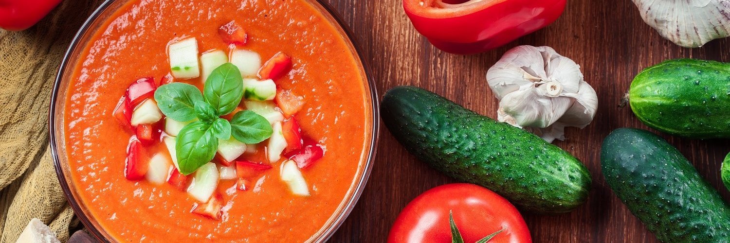 Auf einem dunklen Holztisch steht eine Schale mit roter cremiger Suppe, getoppt mit Gemüsewürfeln und Basilikum. Daneben liegen rote Paprika, Tomaten, Knoblauch und kleine Gurken.