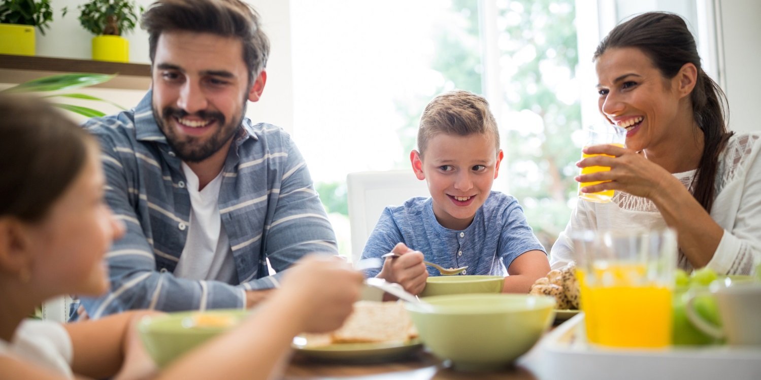 Eine junge Familie mit zwei Kindern sitzt gemeinsam am Tisch beim Frühstück und unterhält sich beim Essen.. Artikel "Essalltag von Familien in belasteten Lebenslagen" öffnen.
