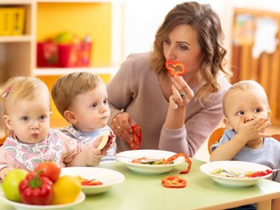 Drei Kleinkinder sitzen am Essenstisch und haben einen gefüllten Teller vor sich. Eine Frau zeigt den Kindern ein Stück Paprika.