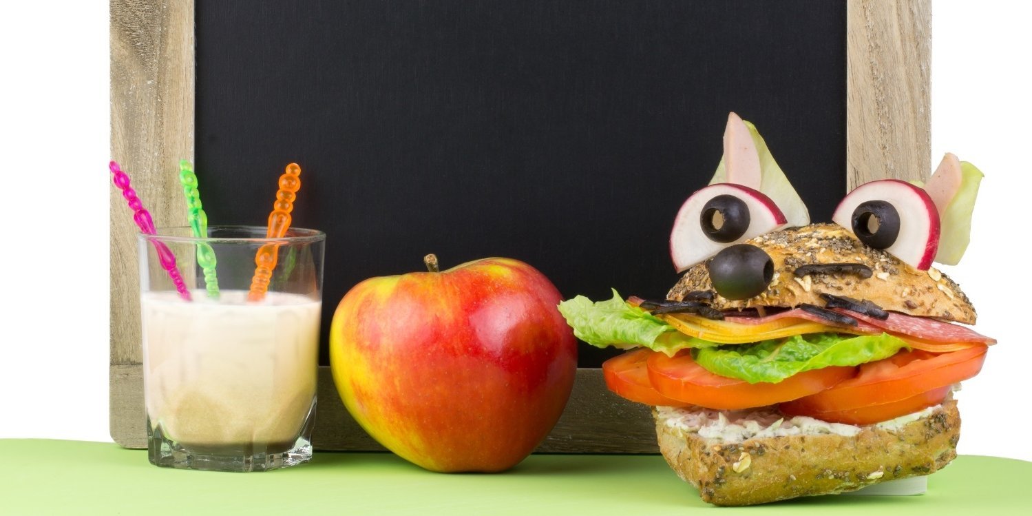 Vor einer kleinen Schultafel stehen auf dem Tisch ein Glas Milch, ein Apfel und ein Vollkornbrötchen mit Salat und Tomate gefüllt und mit Oliven und Radieschen als Fuchskopf dekoriert.. Artikel "EU-Schulprogramm liefert frische Lebensmittel" öffnen.