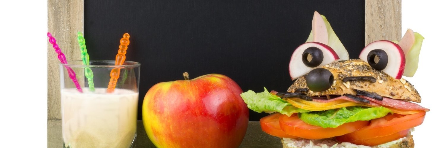 Vor einer kleinen Schultafel stehen auf dem Tisch ein Glas Milch, ein Apfel und ein Vollkornbrötchen mit Salat und Tomate gefüllt und mit Oliven und Radieschen als Fuchskopf dekoriert.