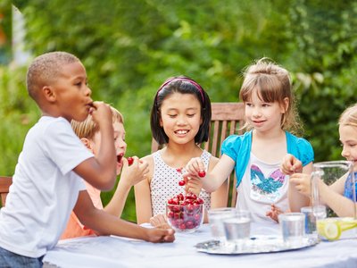 Vier Kinder sind im Freien an einem Tisch, greifen zu Kirschen und essen sie.