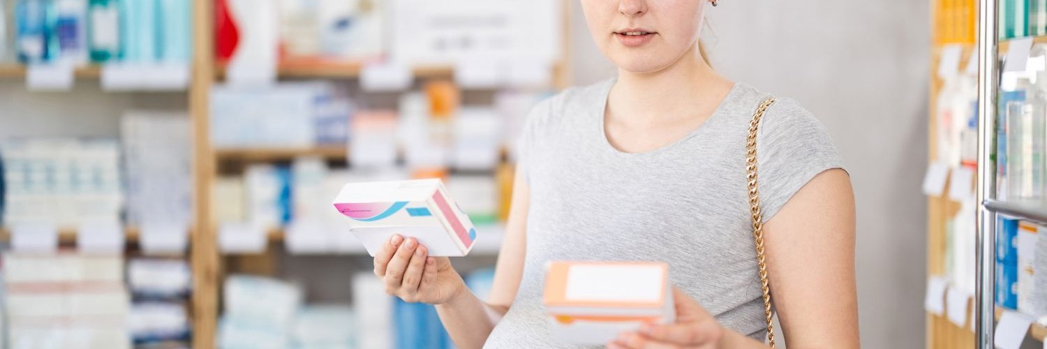 Eine schwangere Frau steht in der Apotheke und hält zwei Verpackungen in den Händen.