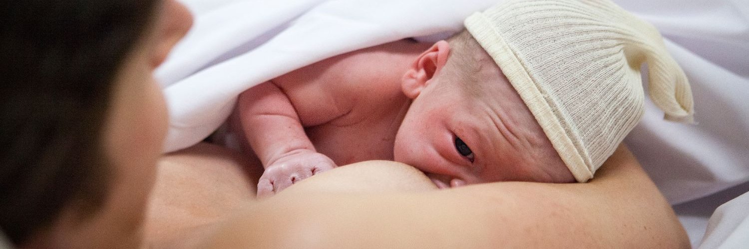 Ein Neugeborenes mit Mütze trinkt an der Brust seiner Mutter, die im Bett liegt.