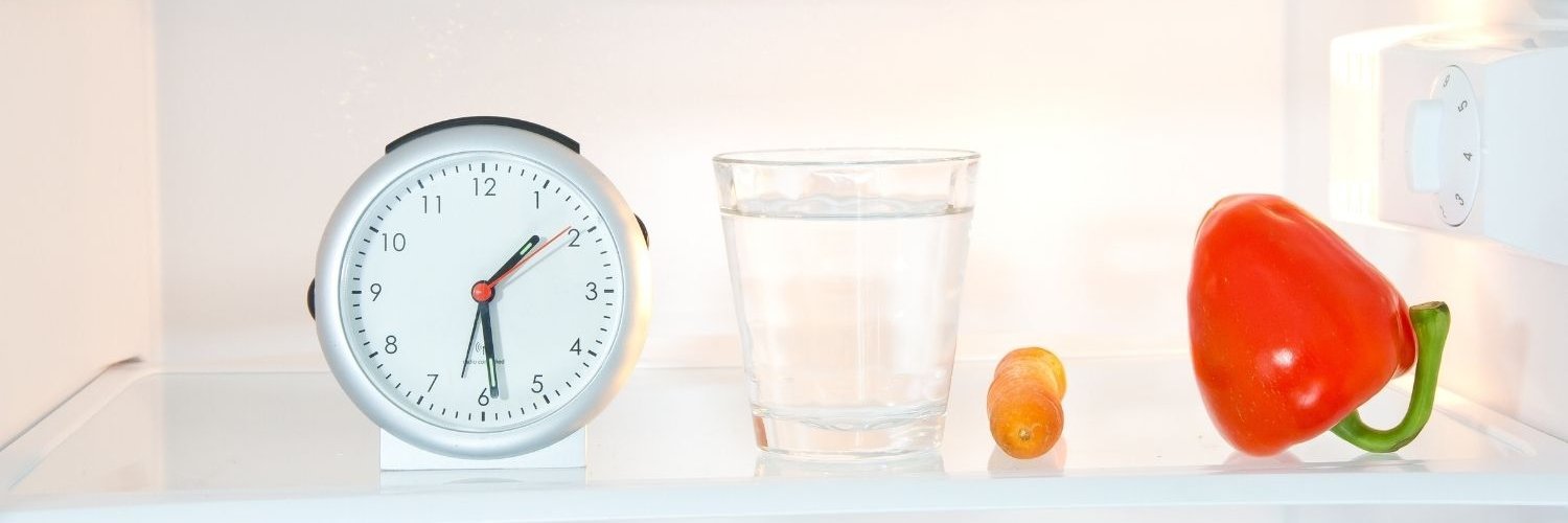 In einem Kühlschrank stehen ein Wecker und ein Glas Wasser, daneben liegen eine Möhre und eine rote Paprika.