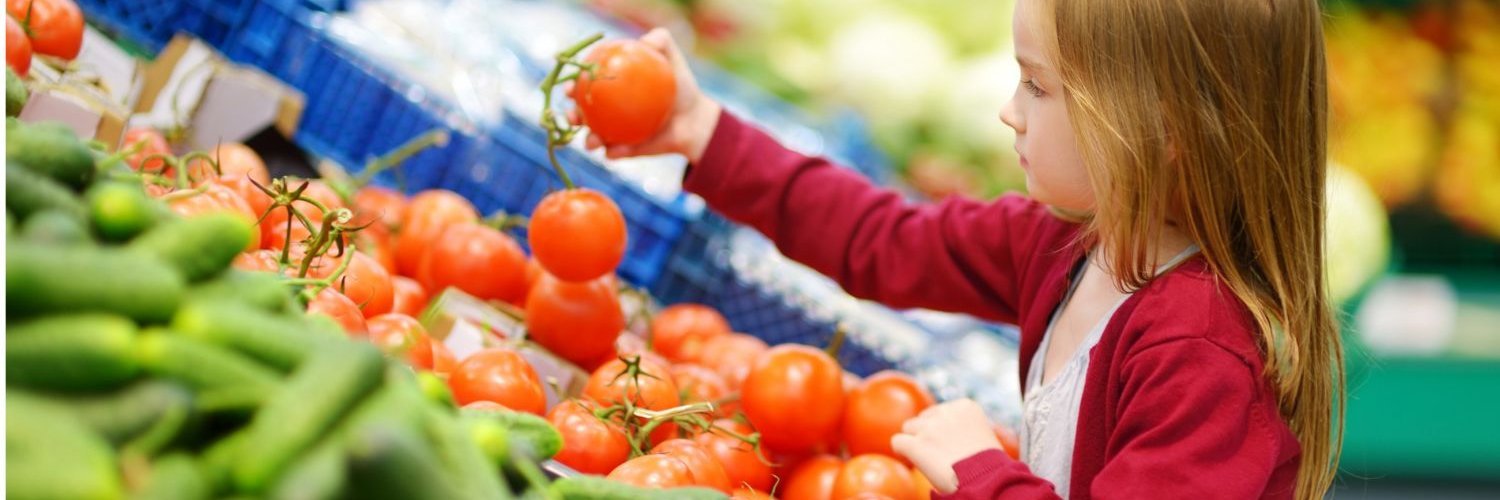 Ein Mädchen im Grundschulalter steht an einer Gemüsetheke im Supermarkt und betrachtet prüfend eine Tomate in ihrer Hand.