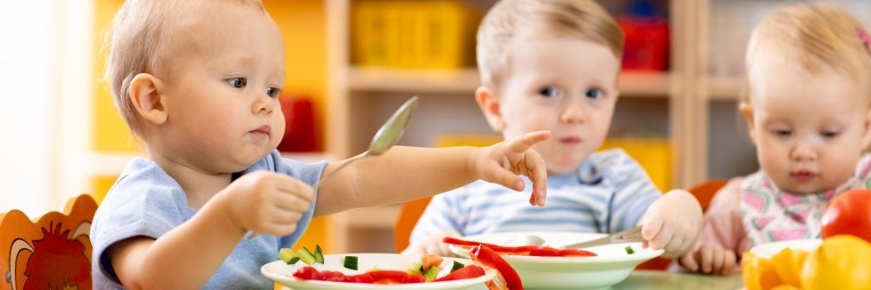 Drei Kleinkinder sitzen mit Tellern am Tisch.