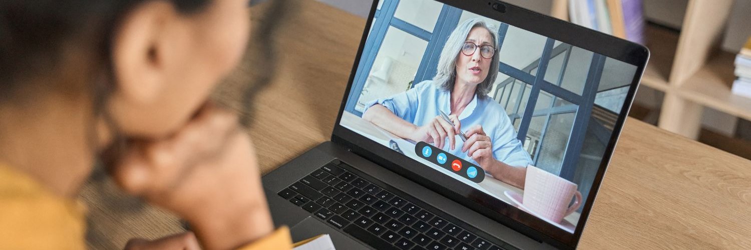 Eine junge dunkelhaarige Frau sitzt vor einem Laptop. Auf dem Bildschirm hält eine ältere grauhaarige Frau einen Vortrag.
