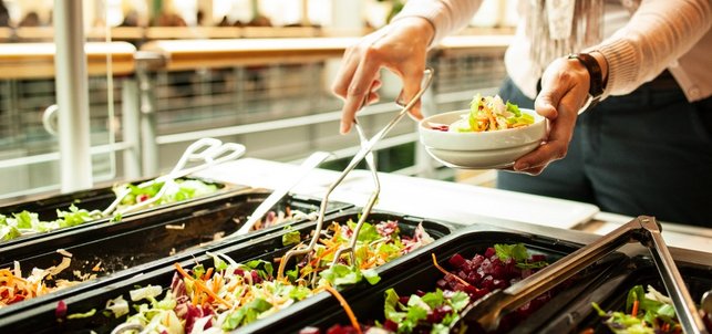 An einer Salatbar in einer Kantine greift eine Person mit einer Küchenzange nach gemischtem Salat, in der anderen Hand hält sie eine kleine Schüssel in der bereits Salat liegt.
