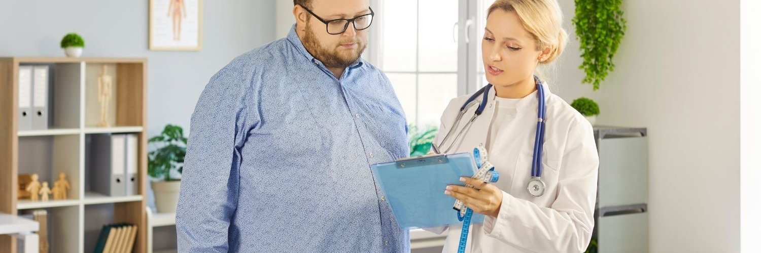 Ein kräftiger Mann im blauen Hemd und eine blonde Ärztin im Kittel schauen gemeinsam auf ein Klemmbrett.
