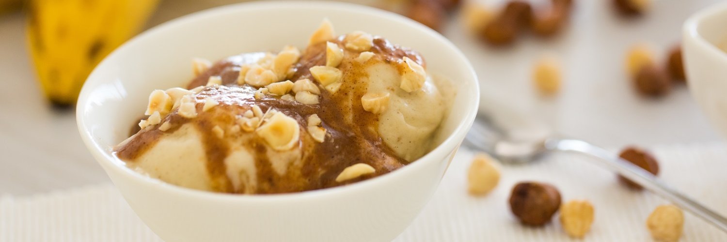 In einer weißen Schale ist Nicecream aus gefrorenen Bananen mit Soße und Nüssen angerichtet. Auf dem Tisch liegen reife Bananen und einige Nüsse.