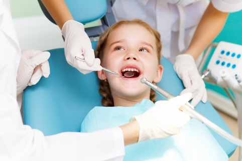 Ein Mädchen sitzt lächelnd auf dem Behandlungs-Stuhl in der Zahnarzt-Praxis. Zwei Personen mit Handschuhen schauen sich die Zähne an.