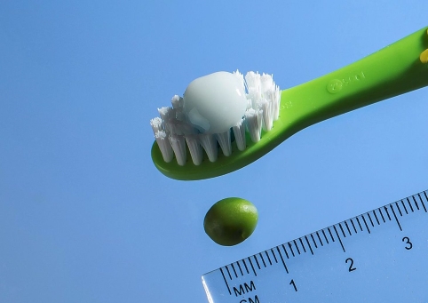 Eine Zahnbürste mit einer Menge Zahnpasta so groß wie eine Erbse, darunter eine Erbse und ein Lineal zum Größen-Vergleich.