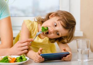 Mädchen hält Handy und wird mit Brokkoli gefüttert