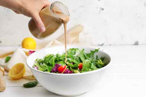 Eine Hand schüttet braune Salatsauce aus einem Glas auf einen Salat.