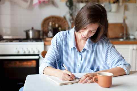 Eine Frau sitzt in der Küche und schreibt etwas in ein Buch.
