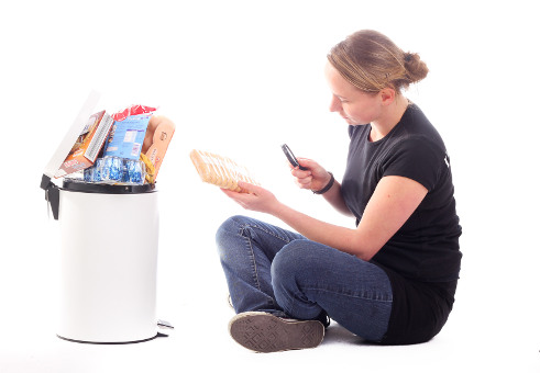Junge Frau sitzt mit Lupe und Lebensmittel vor Mülleimer
