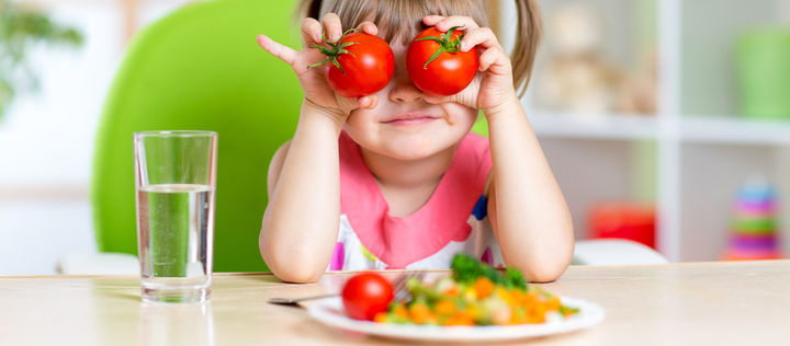 Mächen sitzt vor einem Teller mit Gemüse und hält sich zwei rote Tomaten vor die Augen.
