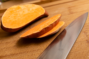 Zwei Scheiben rohe Süßkartoffel vor restlicher Knolle, im Vordergrund eine große Messerklinge.