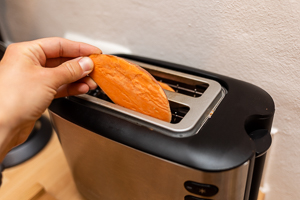 Hand steckt rohe Süßkartoffelscheibe in einen schwarz-silbernen Toaster.