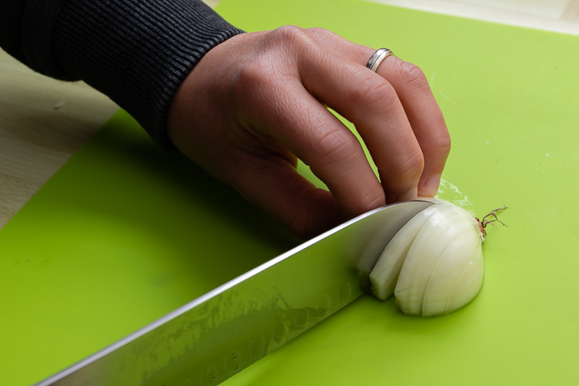 Messer schneidet halbe Zwiebel längs bis zum Strunk ein. Dabei hält eine Hand die Zwiebel im Krallengriff.