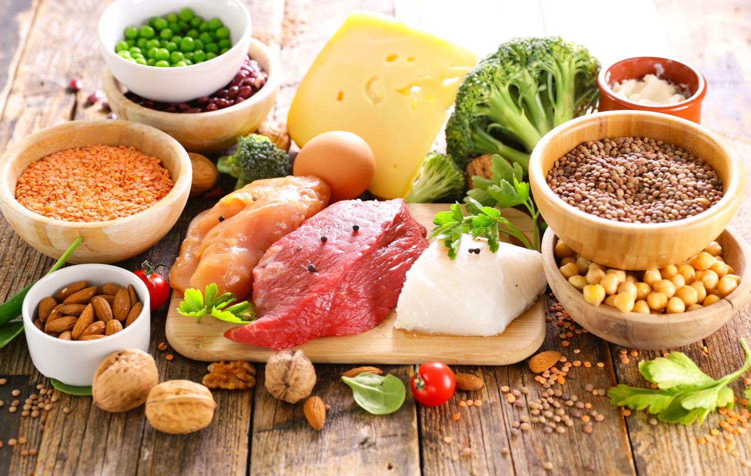 Neben Brett mit Fleisch und Fisch stehen Schälchen mit Nüssen und Hülsenfrüchten und daneben liegen Eier, Brokkoli und Käse.