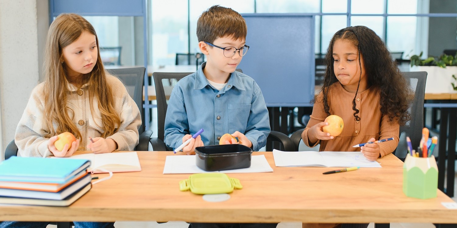 Zwei Mädchen und ein Junge sitzen in der Schule an einem Tisch im Klassenraum. Sie haben ihre Hefte geöffnet, jeweils einen Apfel in der Hand und schauen konzentriert. . Artikel "Mehr Ernährungsbildung in Schulen" öffnen.