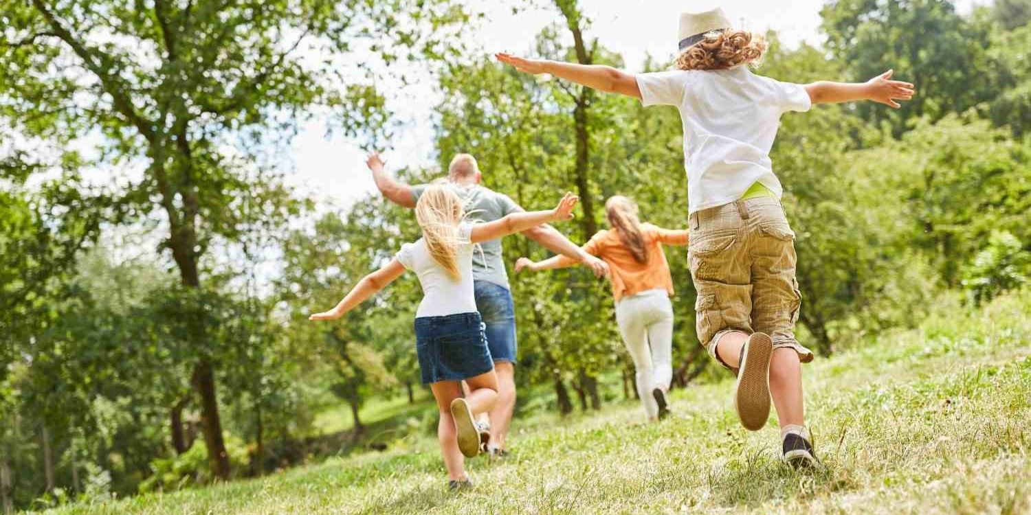 Auf einer Waldlichtung laufen ein Junge und ein Mädchen mit ausgebreiteten Armen hinter ihren Eltern her.. Artikel "Prävention von Adipositas bei Kleinkindern" öffnen.