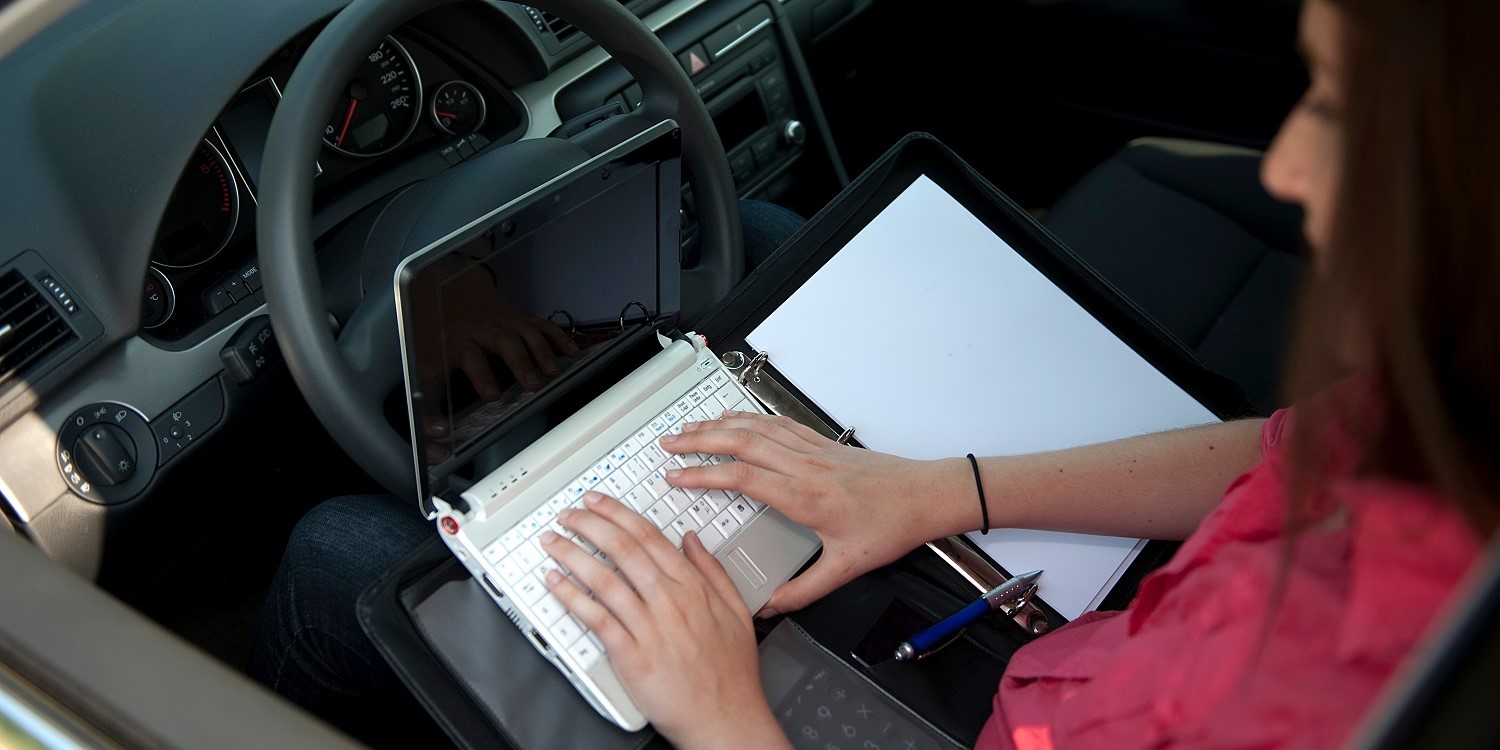 Eine Frau sitzt am Steuer eines Wagens, auf dem Schoß eine Schreibmappe und ein Laptop auf dem sie schreibt. . Artikel "Im Außendienst gut essen " öffnen.
