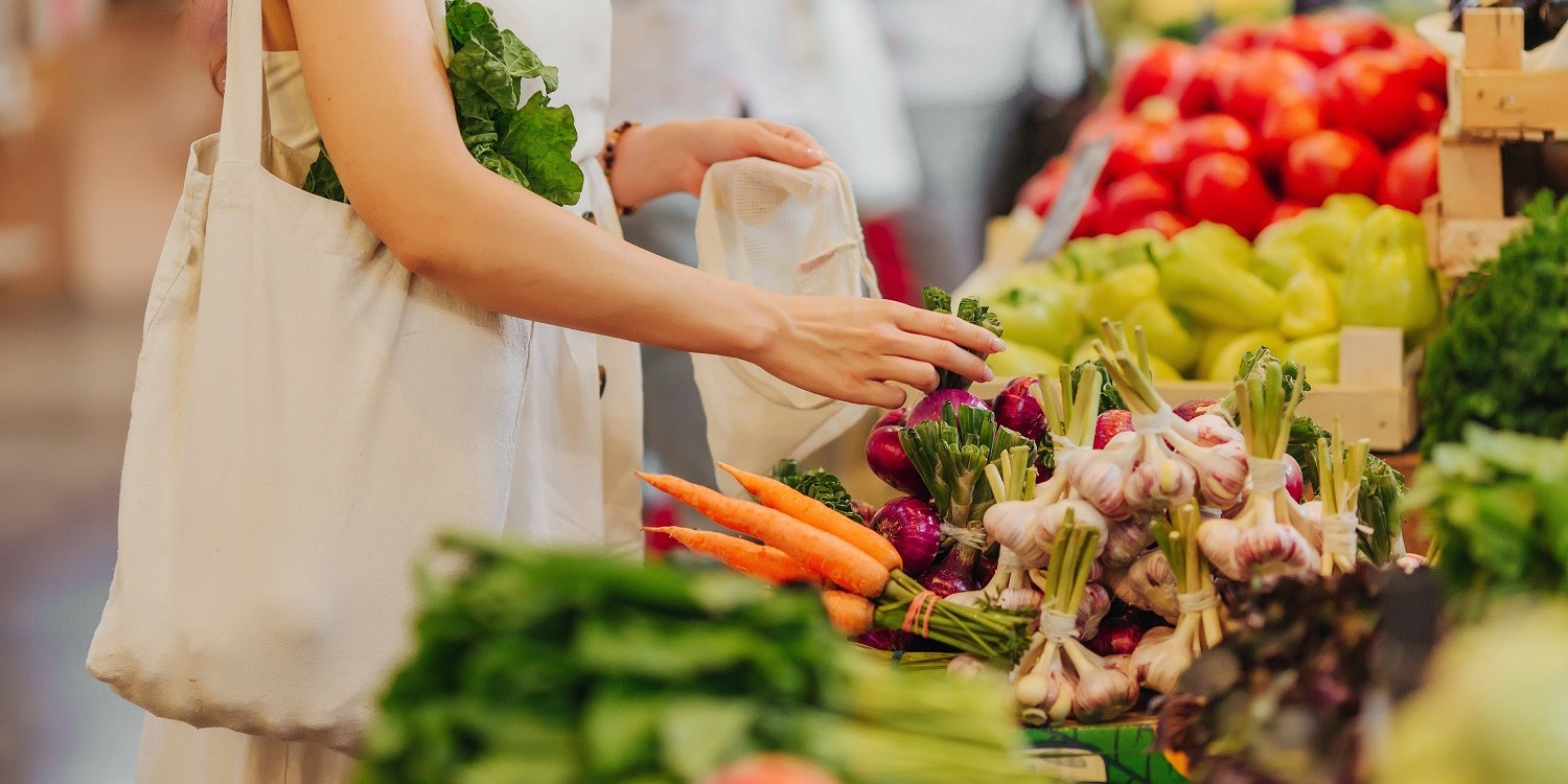 Eine Frau steht auf einem Markt vor einem Obst- und Gemüsestand und packt Gemüse in einen Leinenbeutel. . Artikel "Kaum Pestizidrückstände in Lebensmitteln" öffnen.