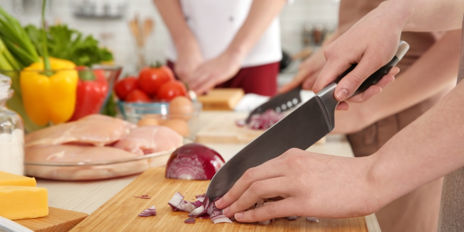 Bei einem Kochkurs schneidet eine Person mit einem großen Messer eine rote Zwiebel auf einem Holzbrett. Im Hintergrund sieht man andere Personen beim Schneiden von Lebensmitteln. . Artikel "Mit Kompetenz vor Ort gegen Ernährungsarmut" öffnen.