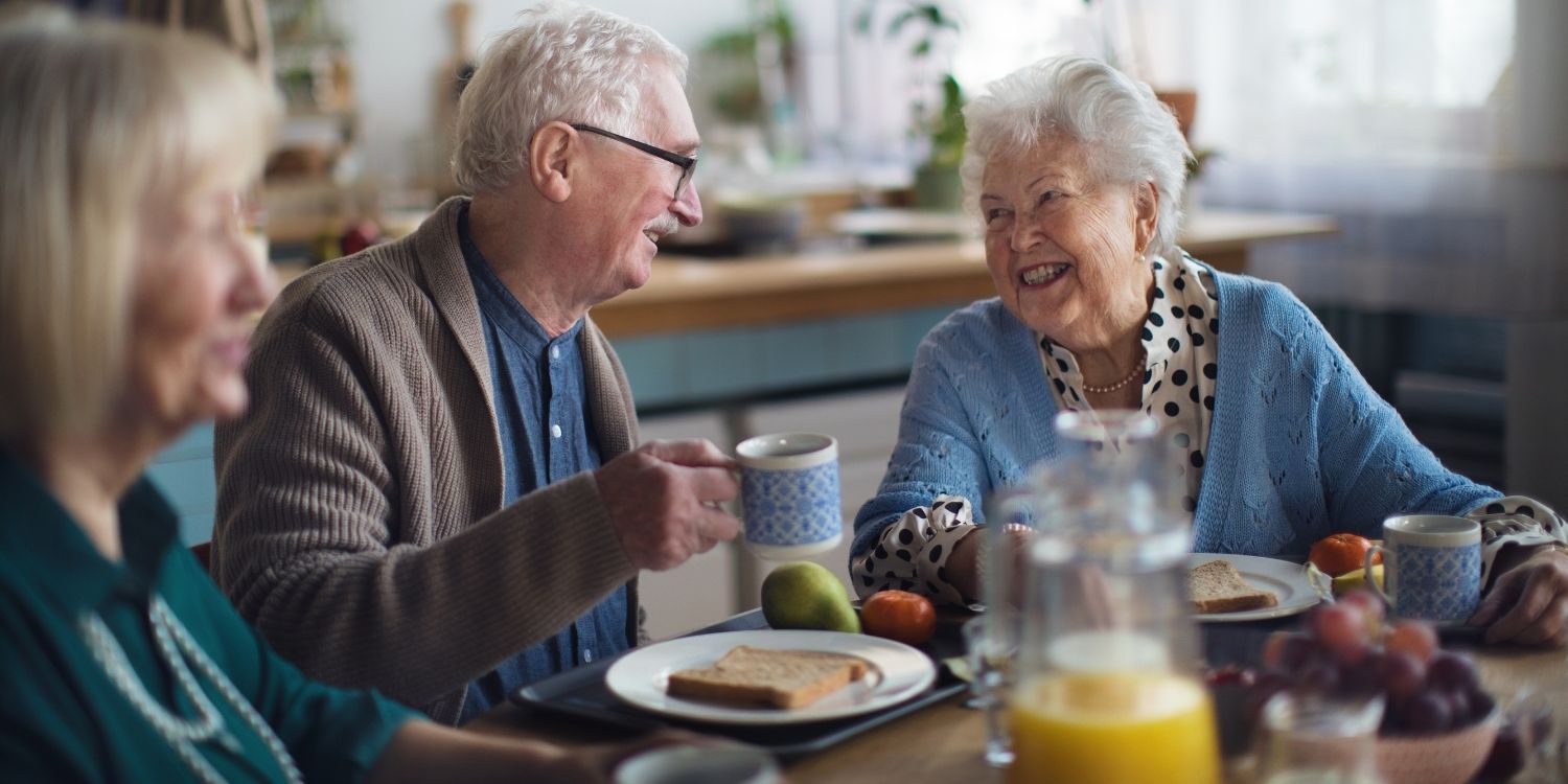 An einem Frühstückstisch sitzen Senioren und Seniorinnen zusammen, eine Frau und ein Mann lächeln einander zu.. Artikel "Gemeinsam essen im Alter" öffnen.