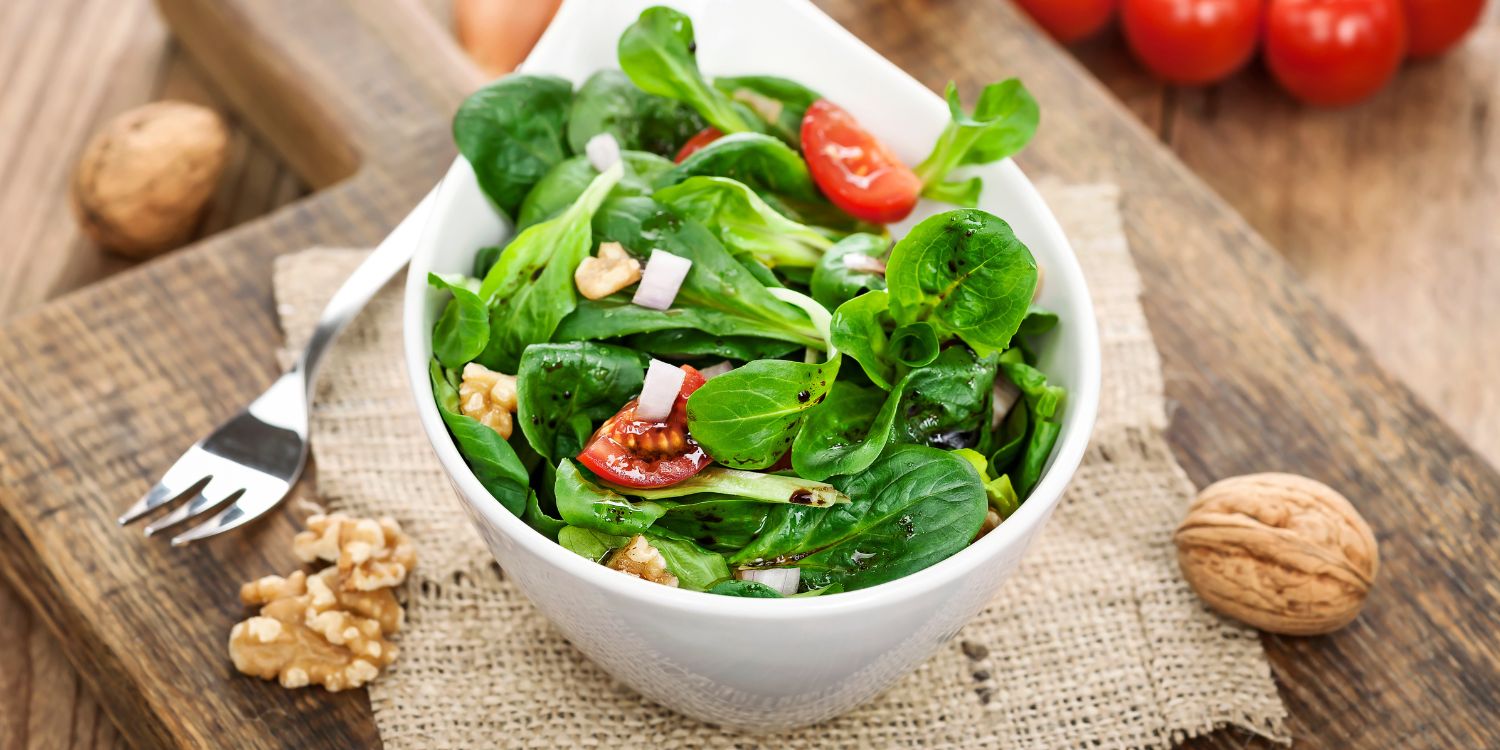 Auf einem Holzbrett steht eine weiße Schüssel mit Feldsalat, Walnüssen, Käsewürfeln und Tomate. Daneben liegen halbierte Walnüsse und eine Gabel.. Artikel "Zeit für frischen Feldsalat" öffnen.