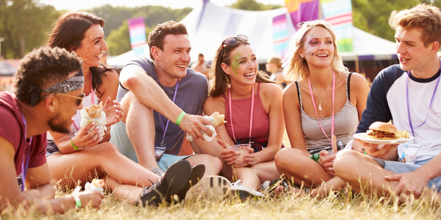 Auf einer Wiese vor einem Veranstaltungszelt sitzen sechs junge Erwachsene im Gras mit belegten Broten, Brötchen oder Schüsseln in den Händen.. Artikel "Fit fürs Festival" öffnen.