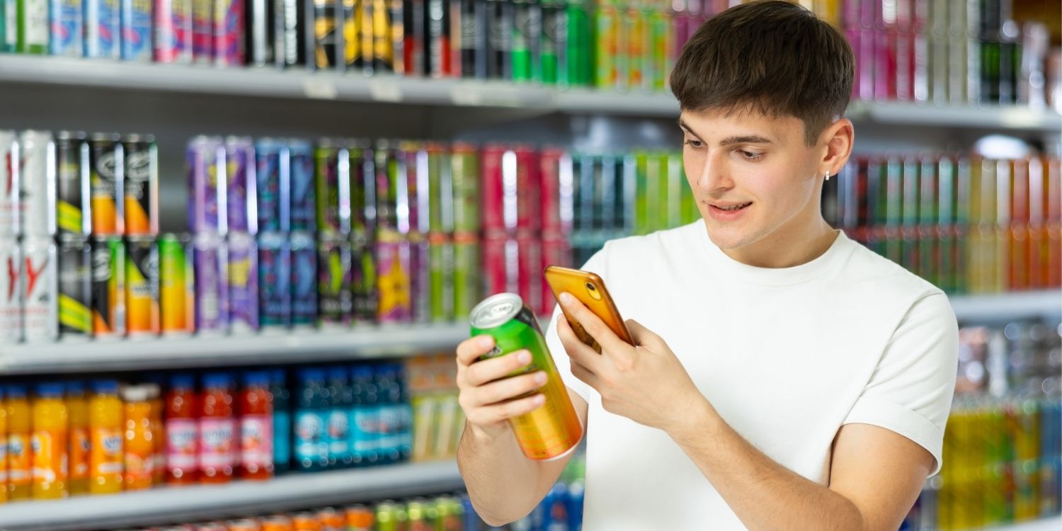 Ein junger Mann steht vor einem Getränkeregal, in der Hand eine Getränkedose und ein Smartphone, auf das er schaut.. Artikel "Funktionelle Limonaden" öffnen.