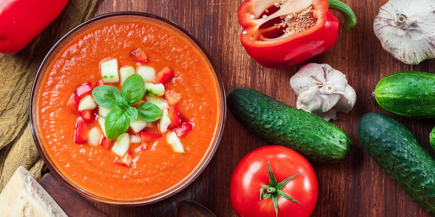 Auf einem dunklen Holztisch steht eine Schale mit roter cremiger Suppe, getoppt mit Gemüsewürfeln und Basilikum. Daneben liegen rote Paprika, Tomaten, Knoblauch und kleine Gurken.. Artikel "Kalte Suppe für heiße Tage: Gazpacho" öffnen.