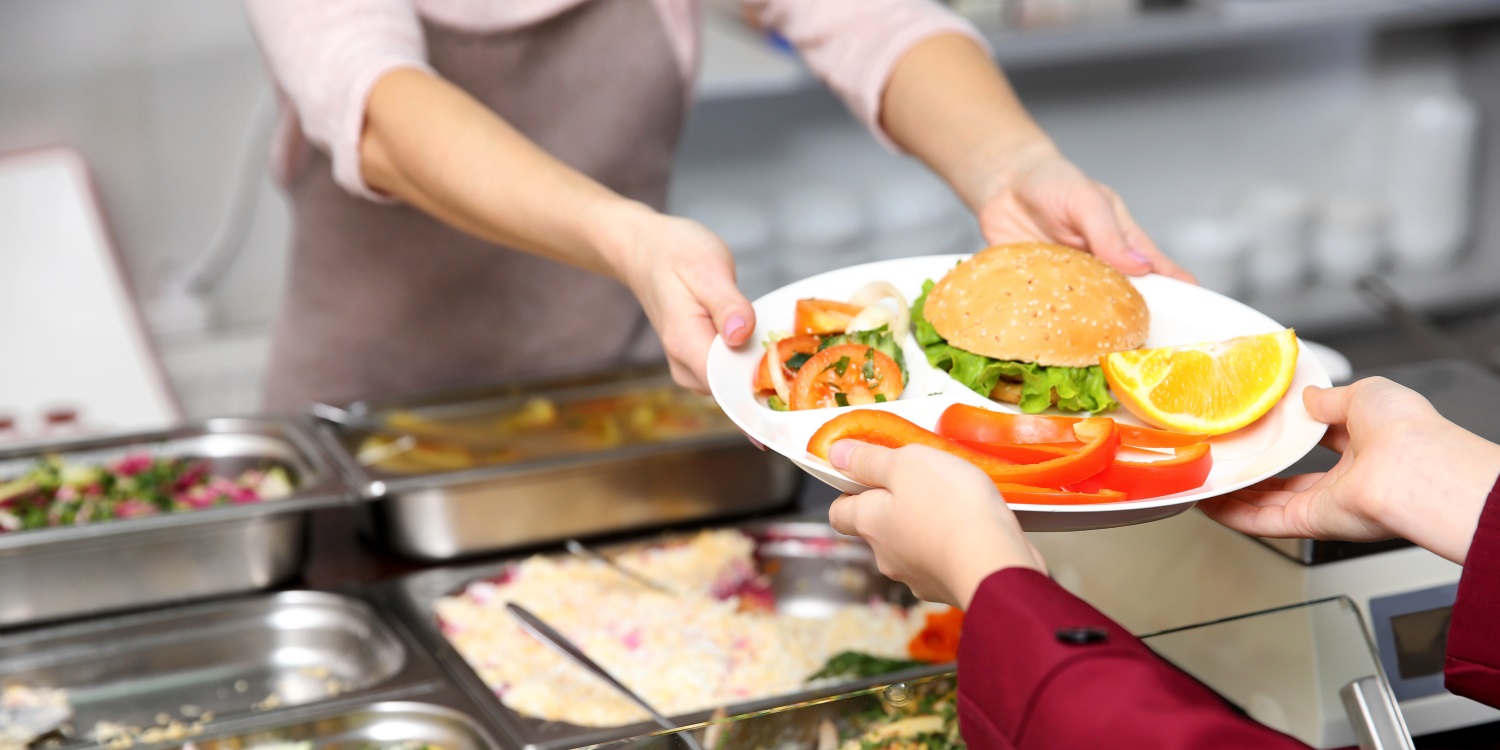 Eine Frau im roten Mantel nimmt an einer Essensausgabe von einer Frau in Schürze einen Teller entgegen, auf dem ein Brötchen, Rohkost, Salat und Obst liegen. . Artikel "Nachhaltige Gemeinschaftsverpflegung stärken" öffnen.