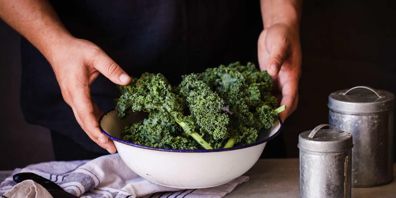 Männerhände stellen eine weiße Schüssel mit Grünkohl auf einen Tisch neben ein Geschirrtuch und zwei Dosen.. Artikel "Grünkohl: Wintergemüse mit Power" öffnen.