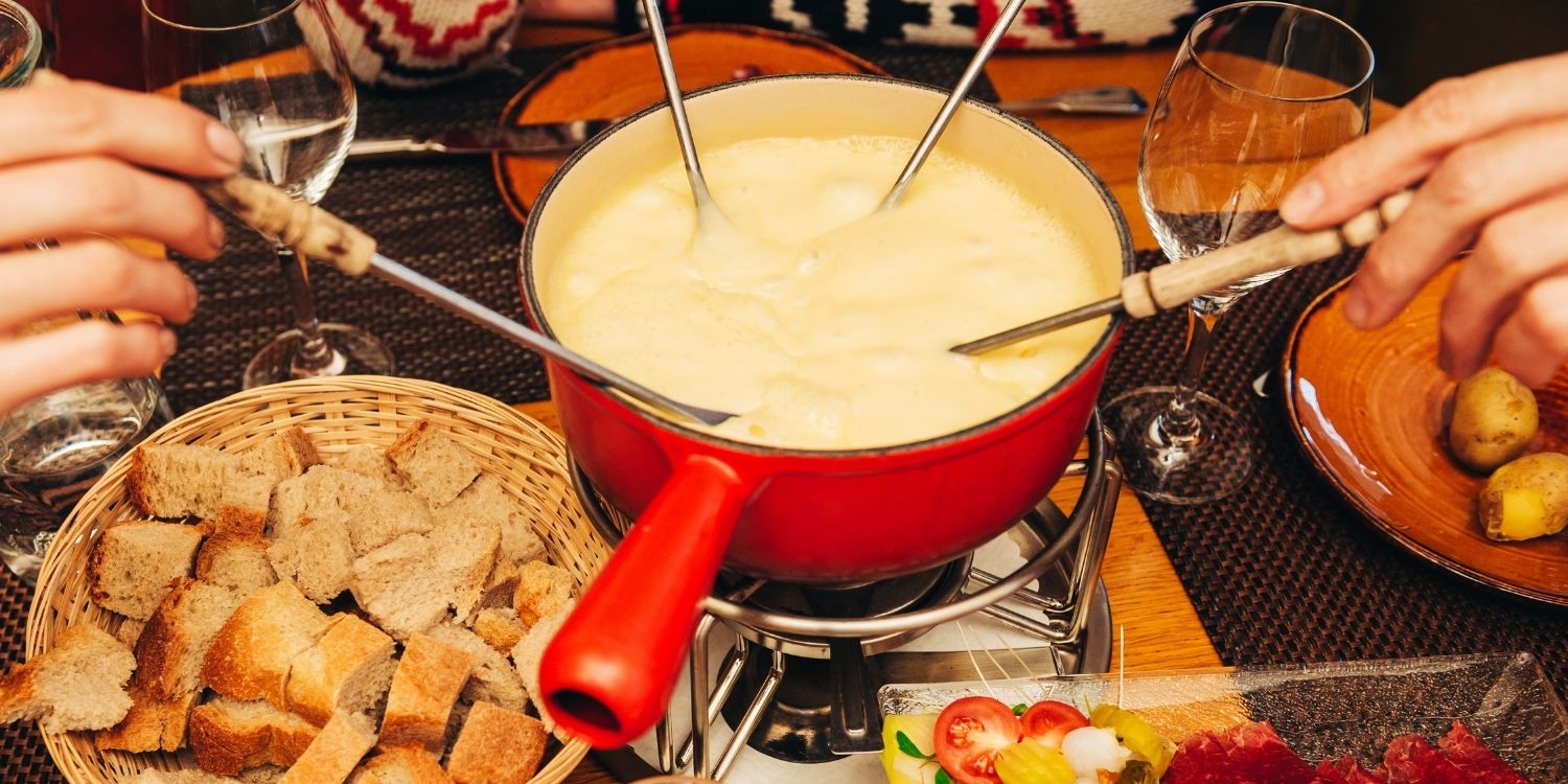 Auf einem Tisch steht eine rote Keramikform mit Käsemasse und vier Fonduegabeln. Ringsum greifen Hände nach den Gabeln und auf dem Tisch steht ein Korb mit Brotstücken, Tomaten und Bündner Fleisch.. Artikel "Ein würziges Käsefondue mit Gemüse" öffnen.