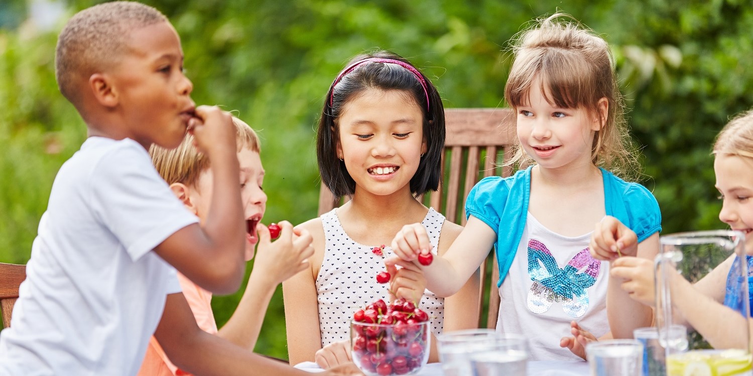 Fünf Kinder im Grundschulalter sitzen draußen gemeinsam an einem Tisch und essen Kirschen. . Artikel "Wege zu gutem Essen für Kinder und Jugendliche" öffnen.