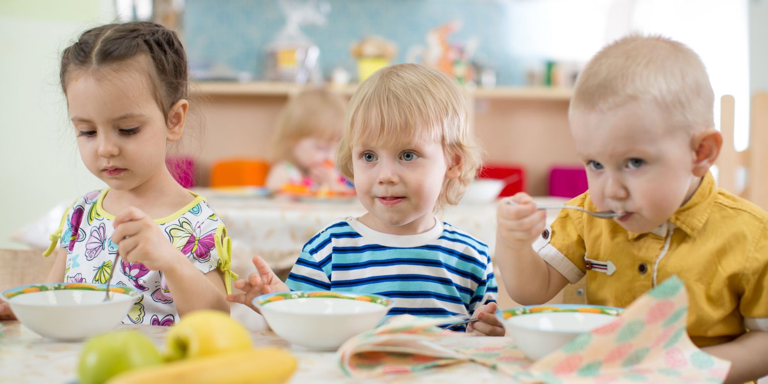 Drei Kleinkinder sitzen in einer Kita beim Essen am Tisch. Vor ihnen stehen tiefe Teller und eine Schale mit Äpfeln und Bananen.. Artikel "So fällt das Mittagessen nicht aus " öffnen.
