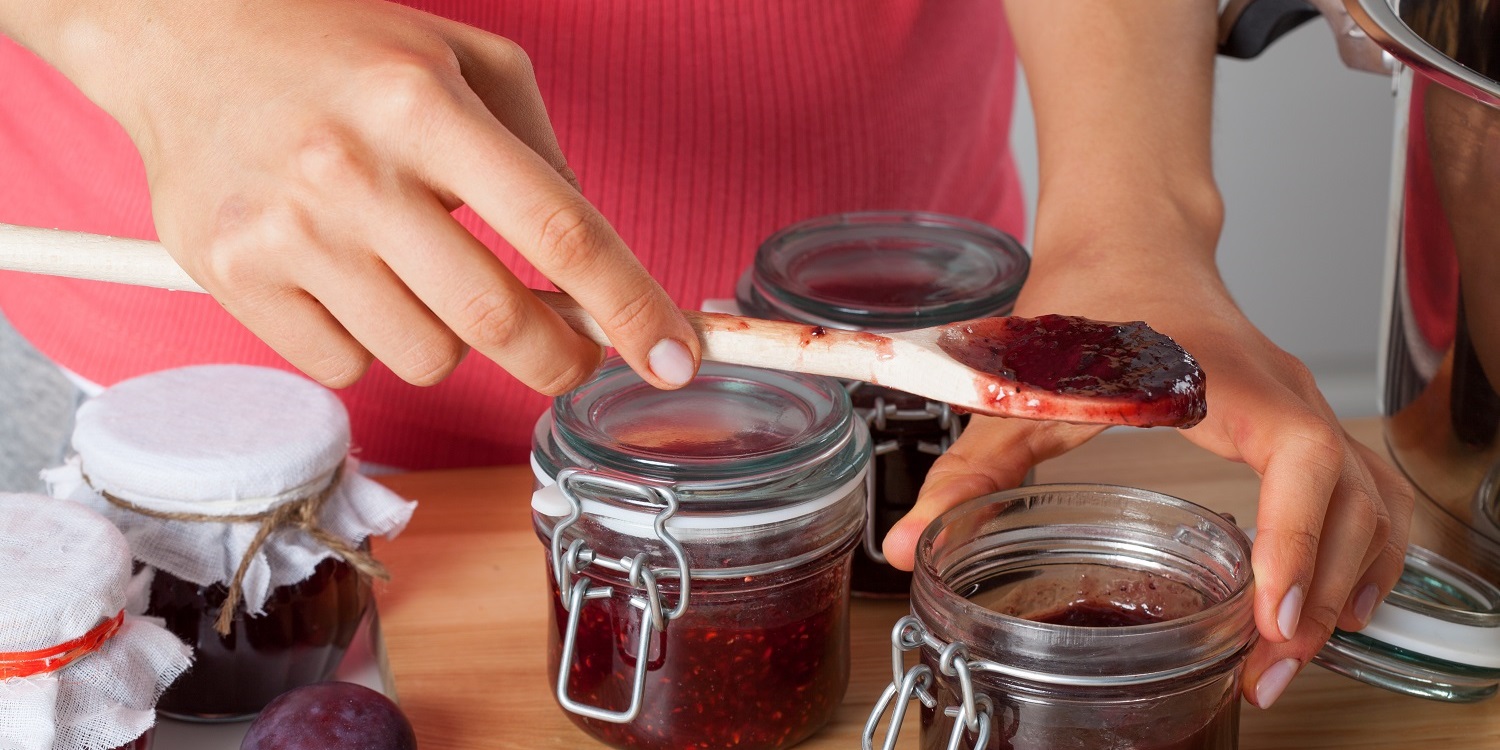 Frauenhände füllen mithilfe eines Holzlöffels Konfitüre aus einem Topf in Gläser. . Artikel "Sommerfrüchte im Glas" öffnen.