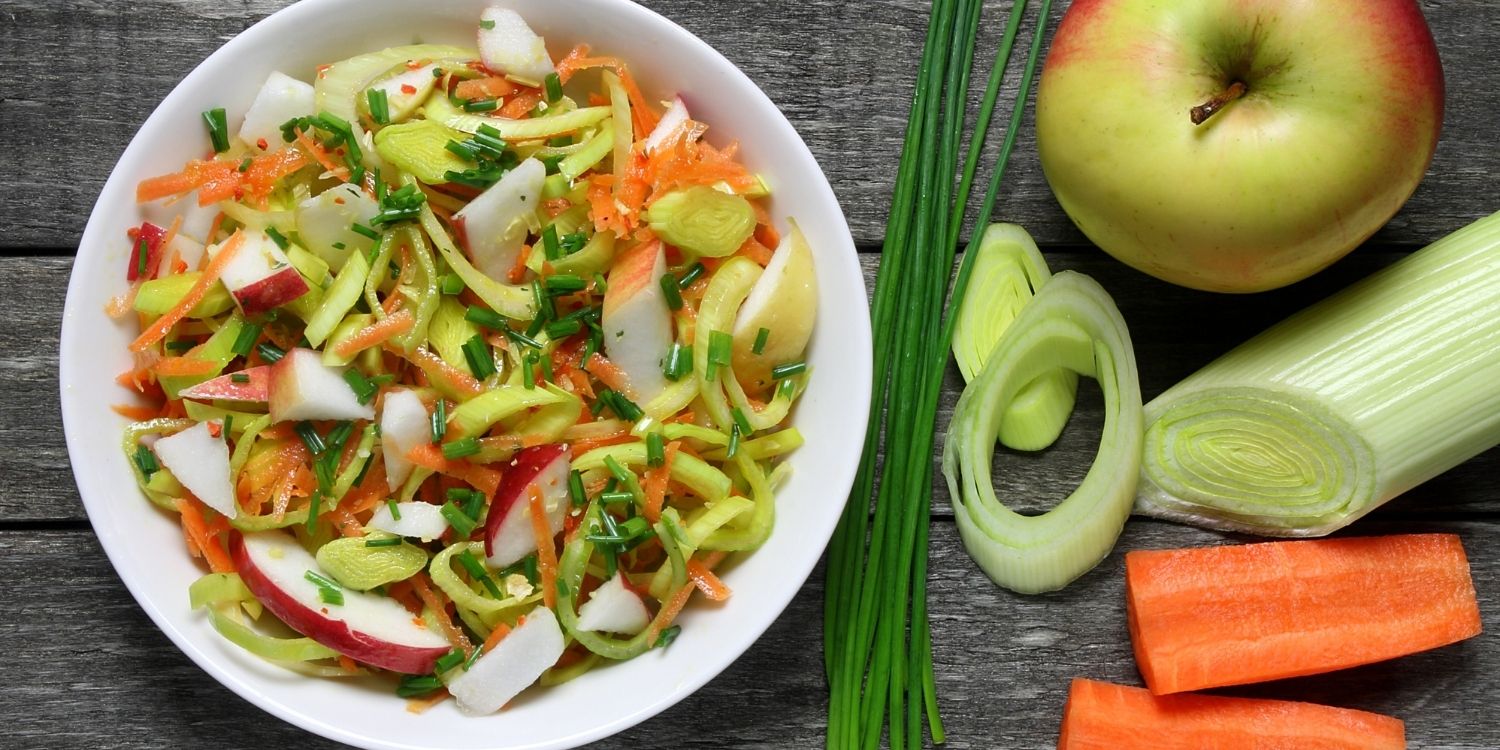 Auf einem dunklen Holzuntergrund steht eine weiße Schüssel mit Salat aus Lauch, Apfel, Möhre und Schnittlauch. Daneben liegen eine frische Lauchstange, ein Apfel, eine halbierte Möhre und Schnittlauch. Artikel "Schnelle Gerichte mit Lauch" öffnen.