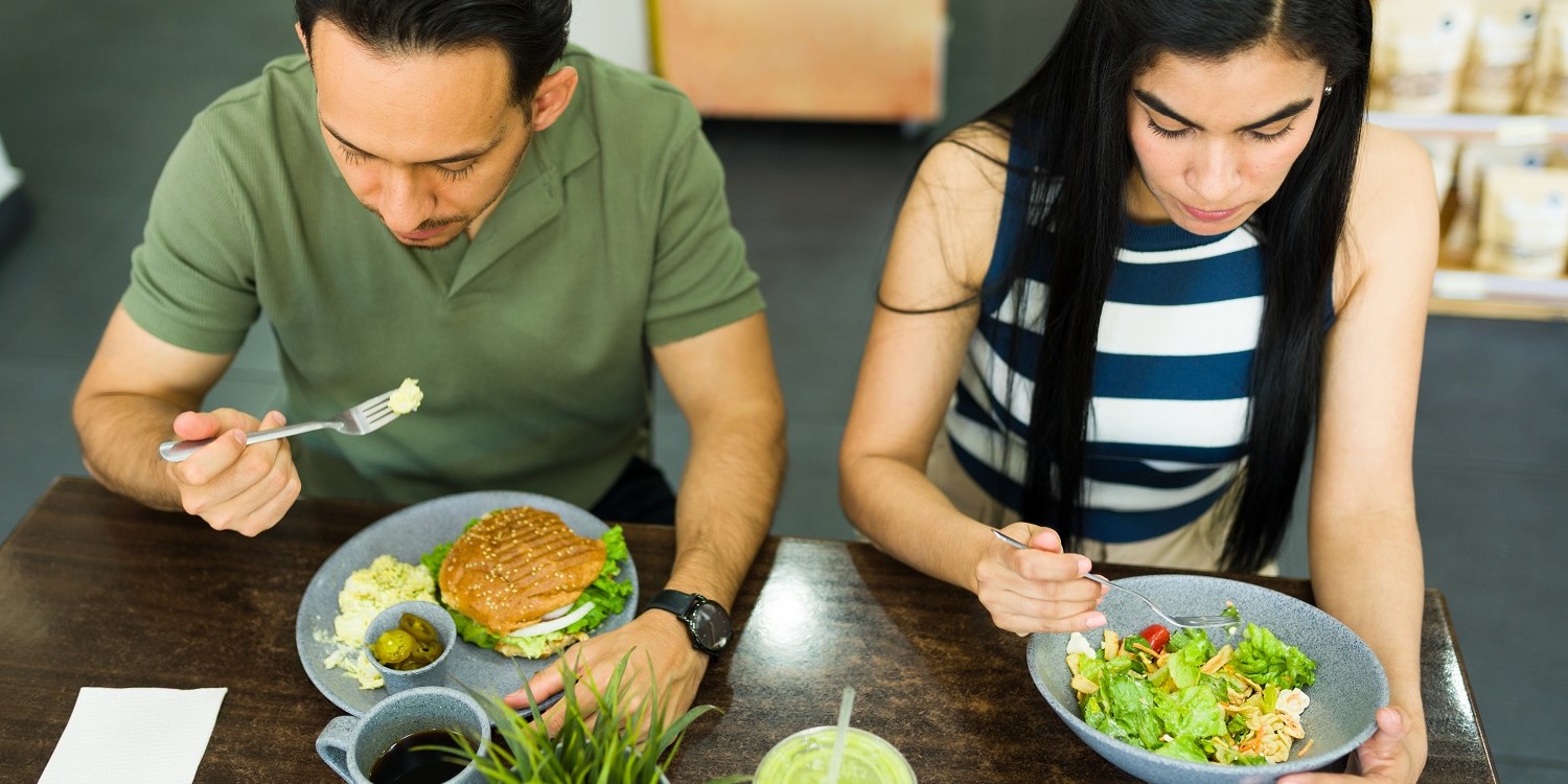 Ein Mann und eine Frau sitzen gemeinsam am Tisch und essen. Er isst einen Burger und sie einen Salat.. Artikel "Fleisch für ihn, Salat für sie " öffnen.