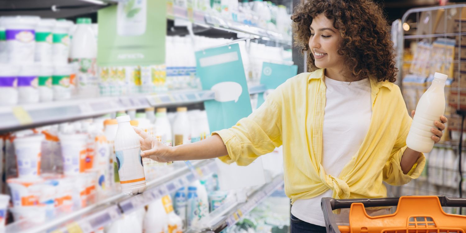 Eine junge dunkelhaarige Frau mit Einkaufswagen hält im Supermarkt eine Flasche Milch in der einen Hand und nimmt mit der anderen ein Milchprodukt aus dem Regal.. Artikel "Frische Fakten rund um Milch" öffnen.