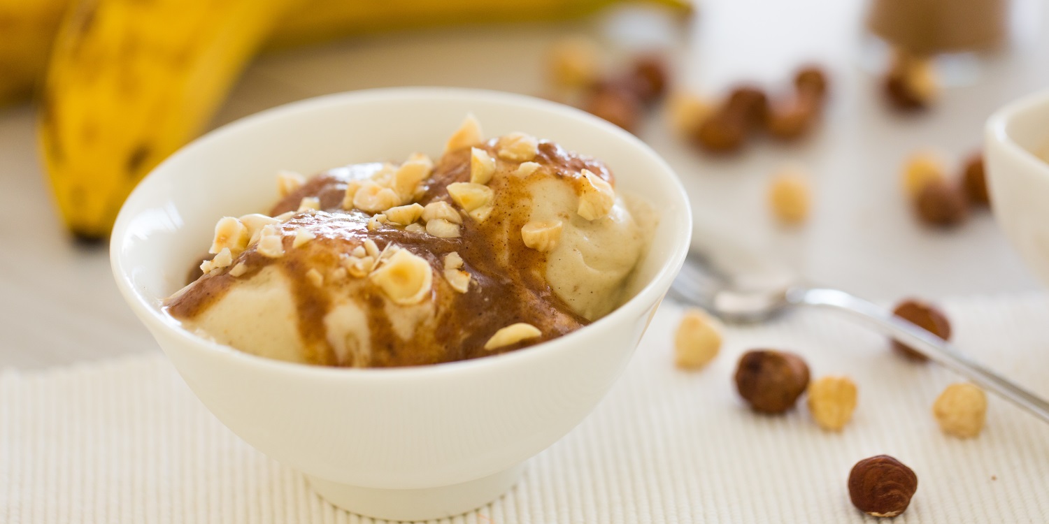 In einer weißen Schale ist Nicecream aus gefrorenen Bananen mit Soße und Nüssen angerichtet. Auf dem Tisch liegen reife Bananen und einige Nüsse.. Artikel "Fruchtige Eiscreme mit weniger Kalorien" öffnen.